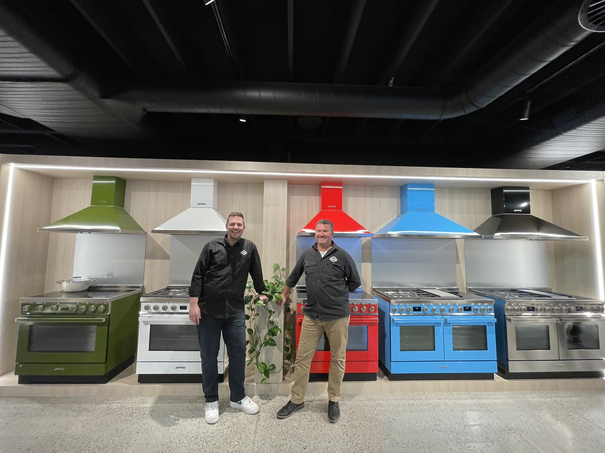 Managing Director Matt Jackson and Andrew Howard of David Reid Homes New England standing in front of a row of Smeg range cookers in green, white, red, blue and stainless with matching rangehoods